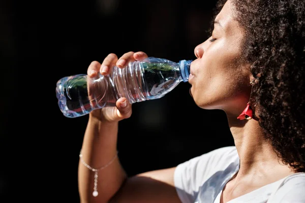 Çekici siyahi kadın sıcak bir yaz gününde bir şişe su içiyor. Doğal hidratlama kavramı.