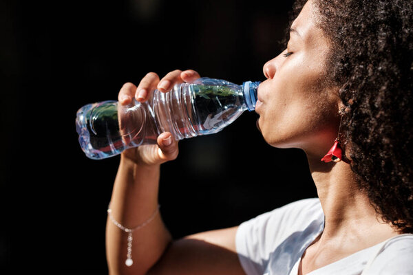 Attractive black woman drinking a bottle os still-water in a hot summer day. Natural hydrating concept.