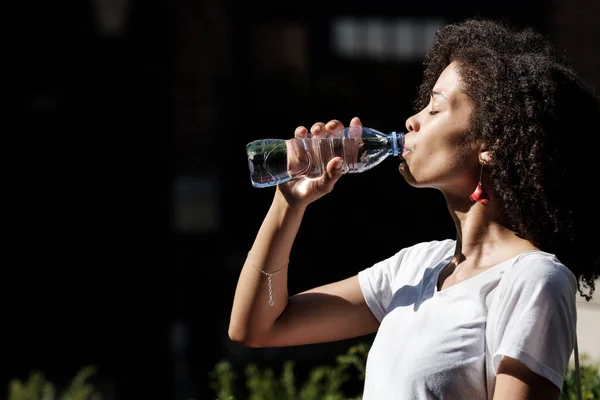 Dışarıdaki şişeden su içen çekici zenci kadın. Sıcak bir yaz günü ve sert bir ışık var. Sıvılaştırma ve sağlık kavramı.