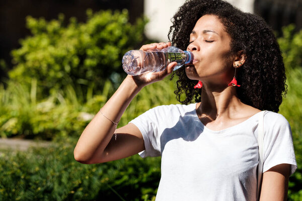 Thirsty curly woman drinking still-water in a park. Her eyes are closed, she is wearing a white t-shirt and red earrings. Health and hydrate concept.