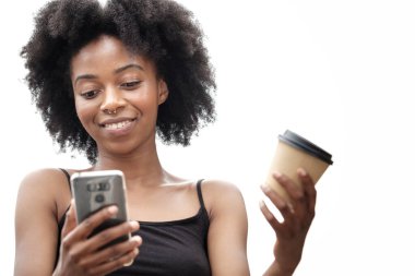 Elinde telefon ve tek kullanımlık kahve tutan gülümseyen bir kadın. Beyaz arka plan. Kıvırcık saçlı ve siyah kolsuz tişörtlü. İletişim ve eğlence kavramı.