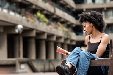 Rahat kıvırcık kadın şehir ortamında bir bankta oturmuş kitap okuyor. Teknoloji dışı yaşam tarzı konsepti. Rahatlayın ve dinlenin..