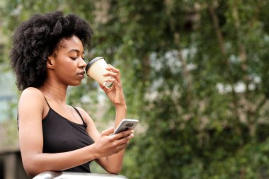 Ciddi siyah kıvırcık kadın kahve içiyor ve telefonda yeşil arka planda daktilo yazıyor. Çalışma molası ve dinlenme konsepti.