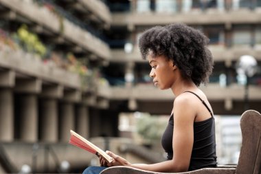 Kıvırcık siyah bir kadın açık hava bankında oturmuş kitap okuyor. Hayal gücü ve rahatlatıcı hobi konsepti. Arkasında gri bulanık bir bina var..