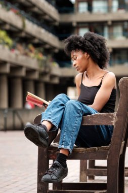 Rahat kıvırcık siyah kadın açık hava bankında oturmuş kitap okuyor. Etrafı gri binalarla çevrili ve çok odaklanmış görünüyor. Eğitim ve edebiyat kavramı.
