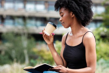 Elinde defter ve kalem tutan kıvırcık siyah kadın tek kullanımlık bir bardaktan kahve içecek. Arka planda bulanık binalar olan bir parkta. Rahatlama hobileri konsepti. Teknoloji içermiyor. Yaratıcılık ve hayal gücü.