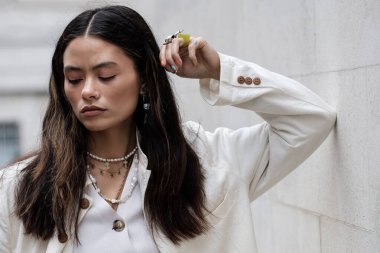 Sokakta beyaz duvara yaslanan dalgın bir kadın manken. Tavır ve karizma gösteriyor. Beyaz bir ceket ve bir sürü mücevher giyiyor..