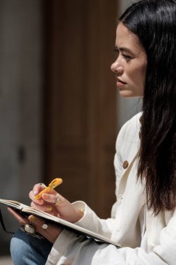 Defter ve kalemle bakan düşünceli kadın. Güneşli bir yaz gününde sokakta oturuyor ve günlüğünü yazıyor..