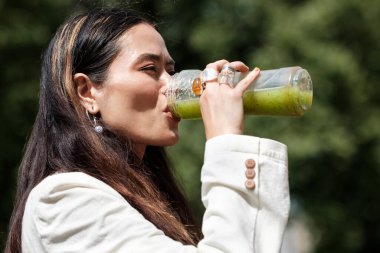 Parkta bir şişe yeşil sağlıklı içecek içen melez bir kadının portresi. Vejetaryen ve sağlıklı vücut konsepti. Doğal yaşam tarzı..