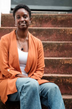 Rahat ve gülümseyen siyah bir kadın Londra 'nın bir caddesinde merdivenlerde oturuyor. Güneşli bir gün ve saçları kıvırcık ve kısa..