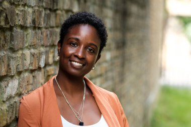 Dışarıdaki tuğladan duvara yaslanan çekici siyahi kadın. Dost canlısı gülümsüyor ve güzel beyaz dişleri var. Turuncu bir ceket, küpe ve tasma takıyor..