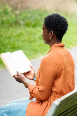 Siyahi bir kadının parkta bir bankta oturmuş kitap okuması. Kıvırcık saçları var. Yaratıcılık ve hayal gücü kavramı. Teknoloji içermiyor.