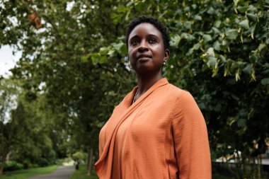 Londra parkında duran dalgın siyah kadın. Turuncu bir ceket giyiyor ve saçları kısa ve kıvırcık. Umut ve gurur kavramı.