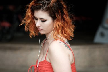 Açık havada müzik dinlemek için telli kulaklık kullanan beyaz kadın portresi. Dövmeler ve kırmızı yaz elbisesi. Saçları kırmızı ve kıvırcık..