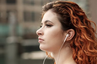 Açık havada müzik dinlemek için telli kulaklık kullanan genç bir kadının vesikalık fotoğrafı. Kafkasyalı, saçları kırmızı ve kıvırcık. Rahatlamış ve rüya gibi bir ifade..