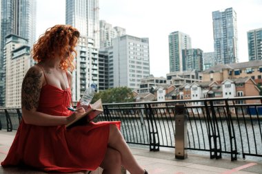 Etrafı gökdelenlerle çevrili beyaz bir kadın defterine kalemle yazıyor. Elinde bir şişe su tutuyor ve kırmızı bir yaz elbisesi giyiyor. Su kanalına yakın..