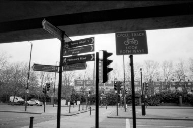 Grainy monochrome analog film photo capturing street signs in london directing to canary wharf, dockmaster's house, hertsmere road, and a cycle track amidst urban buildings clipart