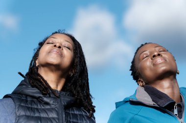 Mavi gökyüzüne karşı iki olgun siyahi kadının rahat portresi. Onlar spor yapan fitness insanları ve güneşli bir sonbahar gününde spor giysileri giyiyorlar. Kısa saç ve sarı saç..