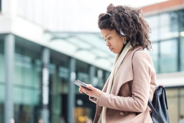Kıvırcık siyah kadın dışarıda elektronik okuyup kulaklık takarak müzik dinliyor. Kış soğuk bir gün ve o ceket giyiyor. Kültür ve klasik hobi kavramı.