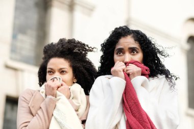 Portrait of two female friends standing the cold outside. They are in an urban environment and they are wearing thick coats and scarfs. clipart