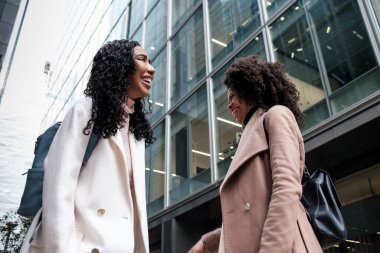 Soğuk bir kış gününde modern binanın önünde gülen iki bayan arkadaş. Kışlık palto ve sırt çantası giyiyorlar. Arkadaşlık ve neşe kavramı.