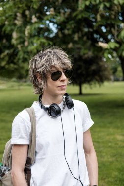 Kulaklık takan ve güneş gözlüğü takan kadın ses mühendisi bir parkta duruyor, su şişesi taşıyan bir sırt çantası taşıyor ve kafasını çeviriyor.