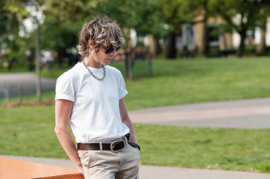 Kısa sarı saçlı, şık bir genç kadın parkta dikiliyor, güneş gözlüğü, beyaz tişört, bej pantolon, kahverengi deri kemer ve gümüş zincirli kolye takıyor.