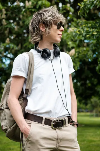 Kulaklık ve güneş gözlüklü şık bir insan. Parkta durmuş, müzikten zevk alıyor ve başka tarafa bakıyor. Günlük giysiler ve sırt çantası takıyor.