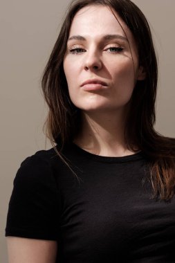 Uzun kahverengi saçlı, sade siyah tişört giyen, doğal ışıkla aydınlatılmış, yüzünde gölgeler ve bej bir arka plan oluşturan ciddi bir genç kadının stüdyo portresi.