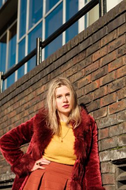 Tuğla duvara karşı poz veren hippi genç bir kadın. Kendine güveni tam görünüyor ve klasik kıyafetler giyiyor. Kırmızı sahte kürk, sarı kazak ve turuncu etek..