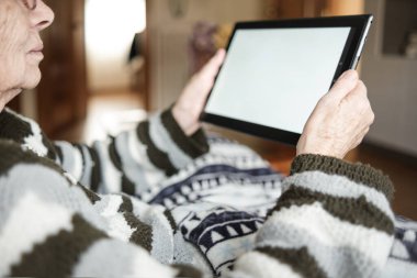 Kanepede oturan ve tablet kullanan tanınmamış yaşlı kadın. Oturma odasında ve battaniye kullanıyor. Teknoloji ve yaşlılar konsepti.