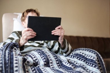 Tablet kullanarak kanepede oturan tanınmamış yaşlı kadın. Yüzü tablet tarafından gizlenmiş ve bir battaniyeyle örtülmüş. Yaşlılar konseptine sahip eğlence ve teknoloji.