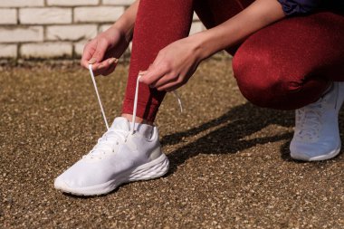 Güneşli bir günde ayakkabı bağcıklarını bağlayan tanınmayan bir spor kadını. Kırmızı taytlar ve beyaz spor ayakkabıları..