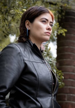 Parkta poz veren çarpıcı bir kadın modelin düşük görüşlü portresi. Uzak ve kendinden emin görünüyor. Ağaçları ve bir sütunu görebiliyoruz..