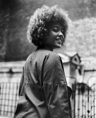 Kameraya bakan, gülümseyen kıvırcık bir genç kadının tek renkli görüntüsü. Bu bir arka plan ve o yürüyor. Şehir titreşimleri, analog film kamerası..