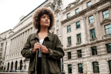 Londra 'nın ünlü bölgesinde, binanın önünde poz veren genç bir kadın model. O, rahat bir şekilde başka tarafa bakıyor. Saçları kıvırcık ve yeşil bir ceketin kenarlarını tutuyor..