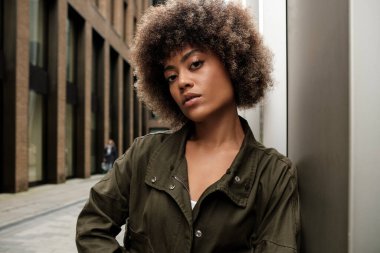 Dar bir sokakta metalik bir yüzeye yaslanan kendine güvenen kıvırcık kadın. Sıradan yeşil ceket giyen melez bir kadın. Arkasında bulanık bir insan var..