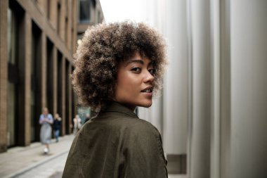 Kameraya bakan genç bir kadın modelin arka görüntüsü. Kıvırcık saçları var ve yeşil bir ceket giyiyor. Arkasında bulanık insanlar var..