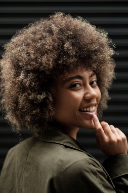 Mutlu genç bir kadın metalin önünde kör bir şekilde poz veriyor ve kameraya bakıyor. Kıvırcık saçlı ve koyu gri ceketli melez bir ırk mankeni..