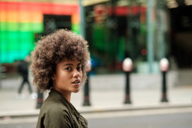 Çekici bir genç kadın renkli ışıklandırma geçmişine sahip kameraya bakıyor. Yolun diğer tarafında bulanık bir kaç tane bulut var. Kıvırcık saçlı.