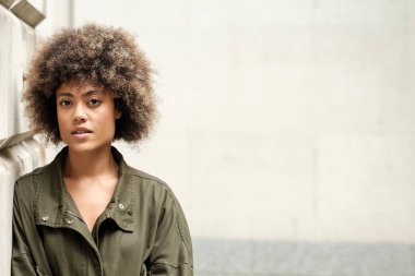 Serene melez kadın dışarıda duvara yaslanıyor. Kıvırcık saçları var ve günlük yeşil bir ceket giyiyor. Kameraya kendinden emin bir şekilde bakıyor..
