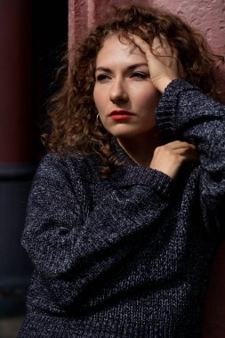 Kafkas kadın portresi beton bir duvara yaslanmış ve başka tarafa bakıyor. Saçları kıvırcık ve gri bir kazak giyiyor. Sol eli saçında..