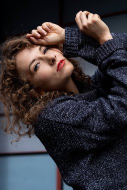 Gri bir arka planı olan, dışarıda poz veren bir model. Saçları kıvırcık ve gri bir kazak giyiyor. Vücudu eğik ve elleri başının etrafında..