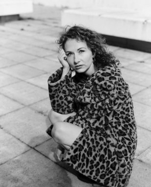 Kendine güvenen bir kadının sahte kürk giydiği ve dışarıda çömelmiş poz verdiği tek renkli bir görüntü. Görüntü bir analog film orta format kamera ile yapıldı. Sahte kürk manto.