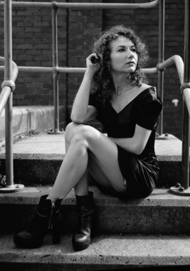 Metalik yapı tırabzanıyla çevrili merdivenlerde oturan kadın model. Siyah bir elbise giyiyor ve başka tarafa bakıyor..