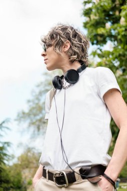 Genç bir insanın portresi güneş gözlüğü takıyor, kulaklık takıyor, beyaz bir tişört giyiyor, parkta duruyor, müzikten ve açık hava atmosferinden zevk alıyor.