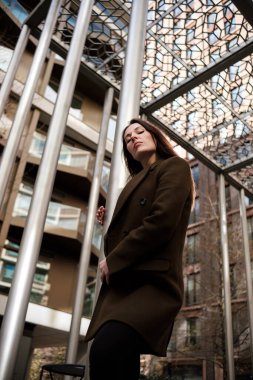 Şehir ortamında modern metal yapının altında poz veren kendine güvenen iş kadınının düşük açılı görüntüsü, çağdaş mimari ve profesyonel tarzı gözler önüne seriyor.