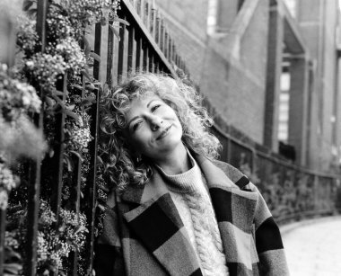 Açık hava duvarına yaslanmış gülümseyen bir kadının rahat portresi. O bir ekose ceket giyiyor ve onun saçı kıvırcık. Monokrom film analog portresi.