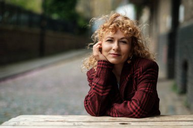 Sokaktaki ahşap masada oturan ve kameraya bakan otantik bir kadın. O, kırmızı bir ekose ceket giyiyor ve kafası sağ eline dayanıyor. Yumuşak ışık ve rüzgar.