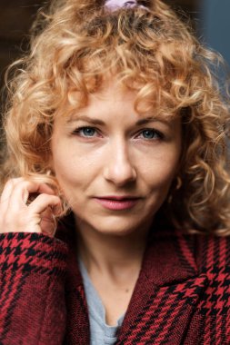 Dışarıdaki kameraya bakan kıvırcık kadının güçlü bir portresi. Kırmızı bir ekose ceket giyiyor ve sağ eli kızıl saçlarına dokunuyor..